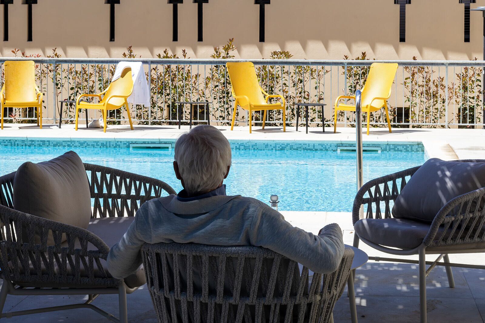 Una persona gran asseguda al costat d'una cadira relaxant a la piscina, amb una piscina i una filera de cadires grogues al fons.