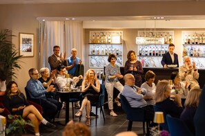 A group of people seated at tables in a room with a bar area in the background. - Guild Siena Senior Resort & Residence (Siena)