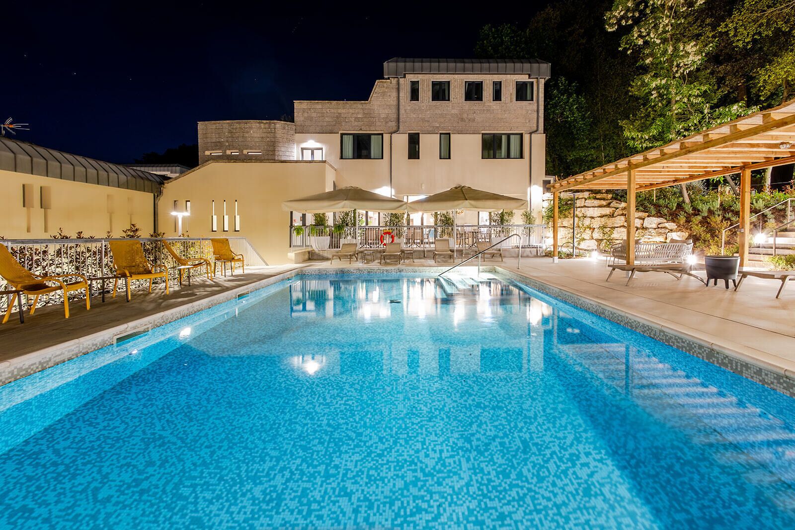 A swimming pool with lounge chairs and umbrellas, surrounded by a building with a stone facade and a covered outdoor seating area.