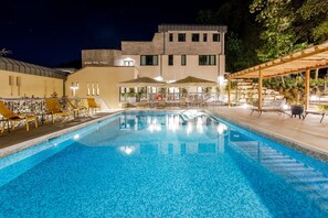 A swimming pool with lounge chairs and umbrellas, surrounded by a building with a stone facade and a covered outdoor seating area. - Guild Living Siena Senior Resort & Residence (Siena)
