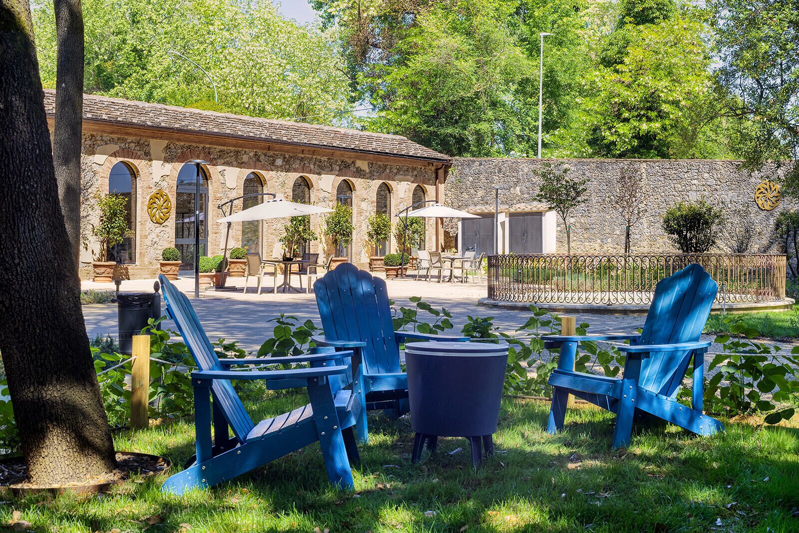 Outdoor seating with chairs and tables, a stone building with arches, and a stone wall in the background.