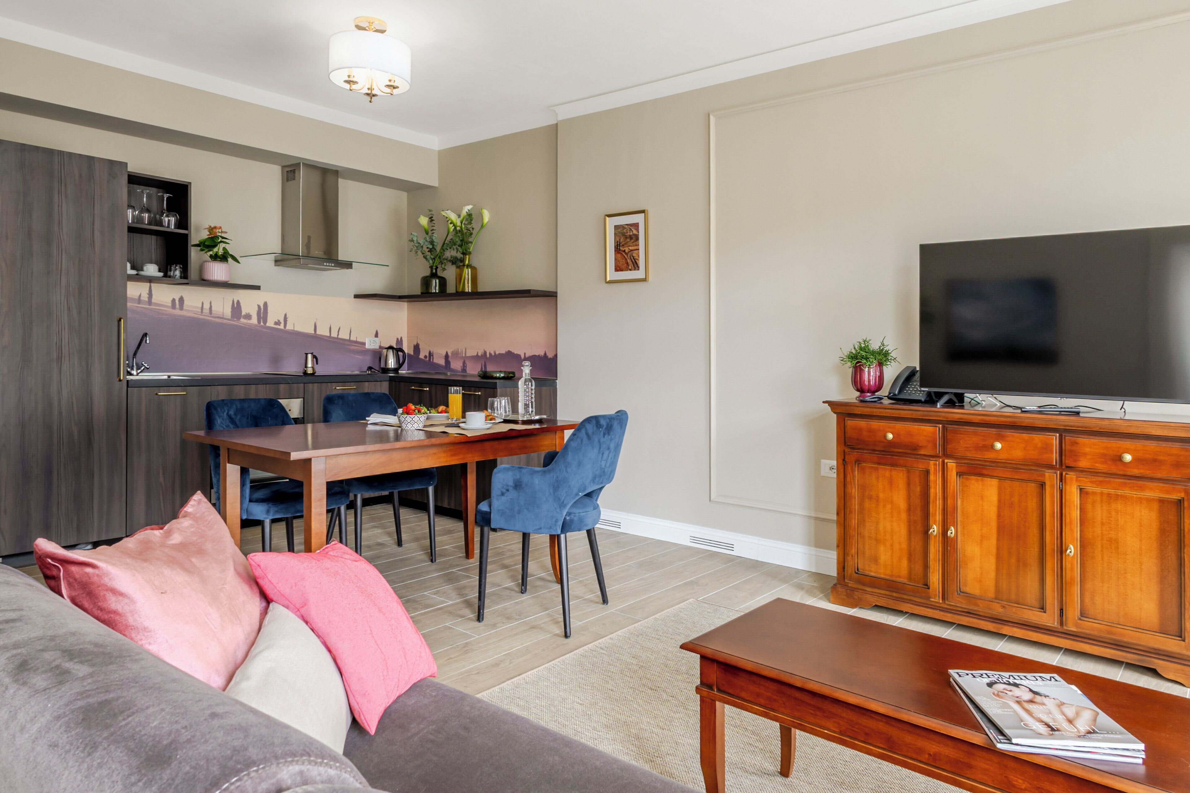 A modern living room with a sofa, a coffee table, a TV, a dining area with chairs, and a kitchen with a stove.