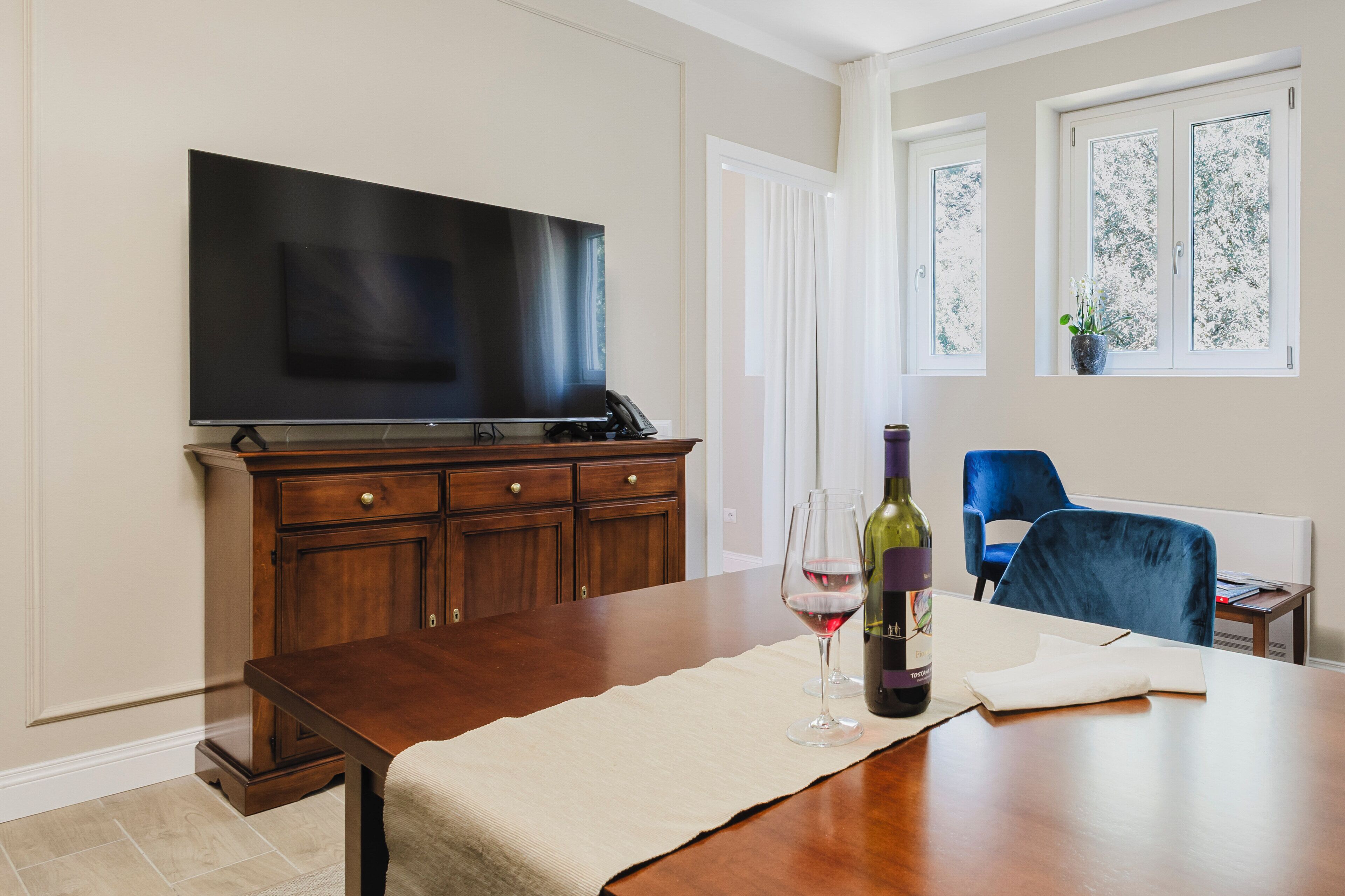 Una sala de estar con televisor de pantalla plana, un mueble de madera, una mesa de comedor con posavasos, copa de vino y botella, una silla azul, y una ventana con planta.