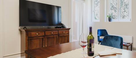 Una sala de estar con televisor de pantalla plana, un mueble de madera, una mesa de comedor con posavasos, copa de vino y botella, una silla azul, y una ventana con planta.