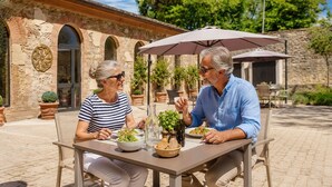 An outdoor patio with stone walls, two people dining at a table with food and drinks, and an umbrella providing shade. - Guild Living Siena Senior Resort & Residence (Siena)