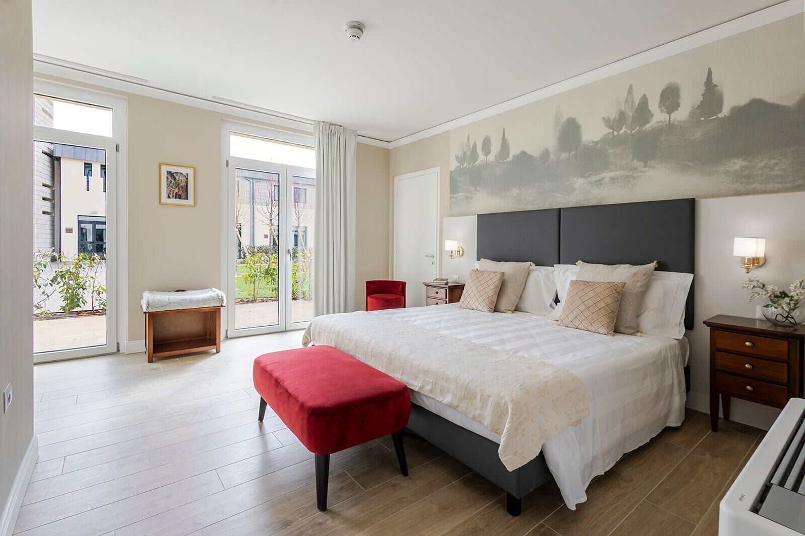 A bedroom with a large bed, a bench, a nightstand, a lamp, and a painting on the wall.