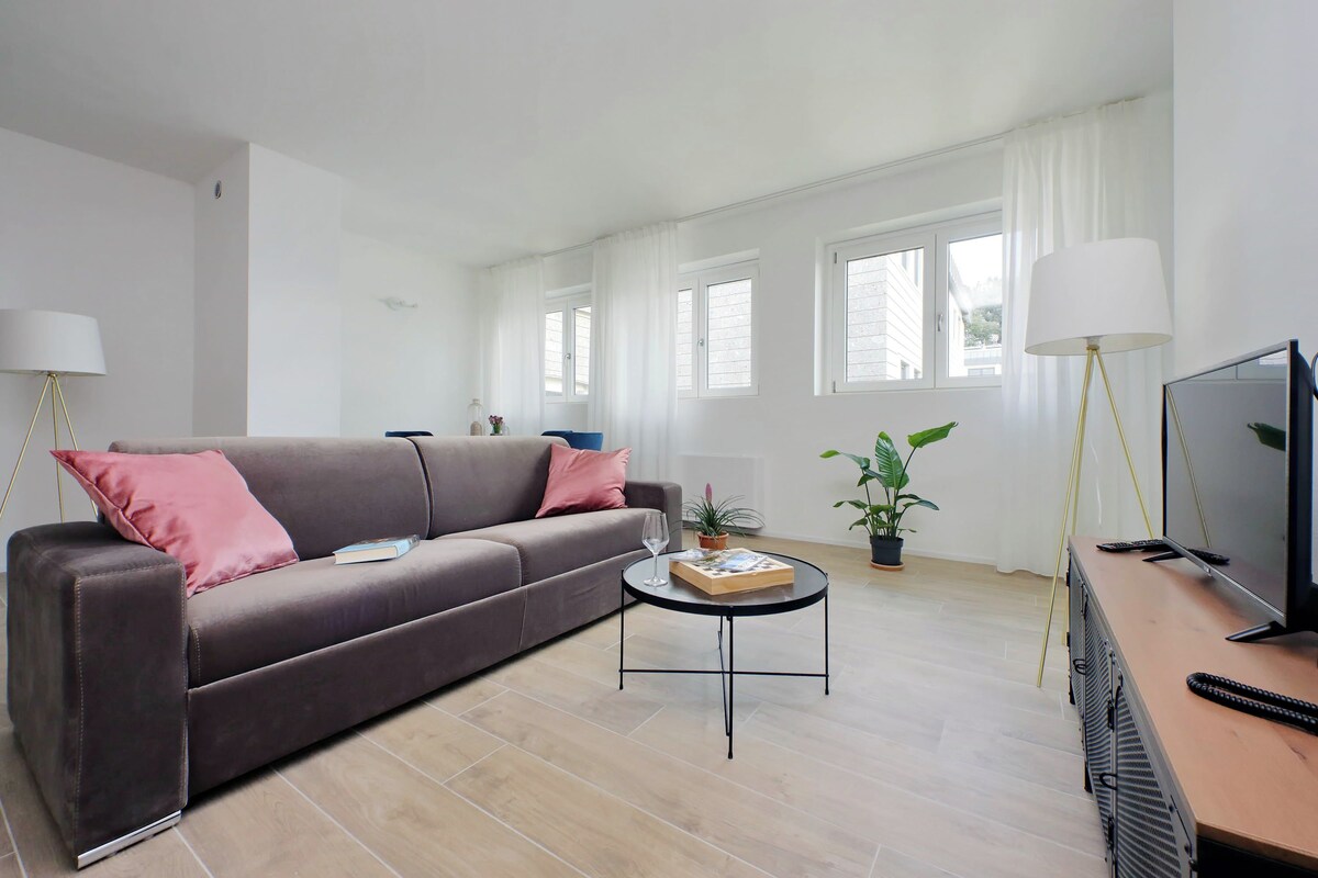 A modern living room with a grey sofa, a coffee table with a book and a glass, and a TV on a wooden stand.