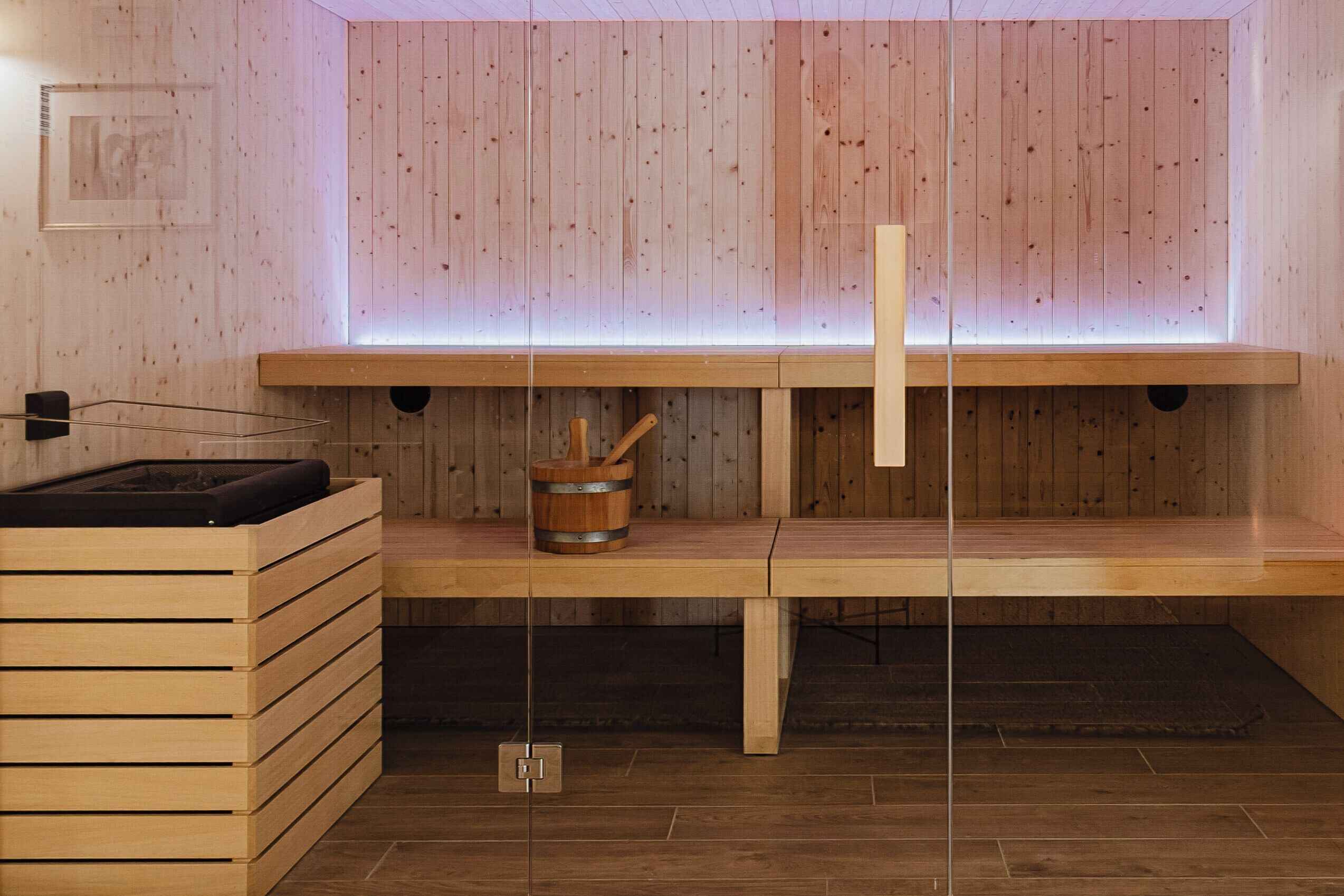 A wooden sauna with a bench, a wooden bucket, and a wall-mounted light fixture.