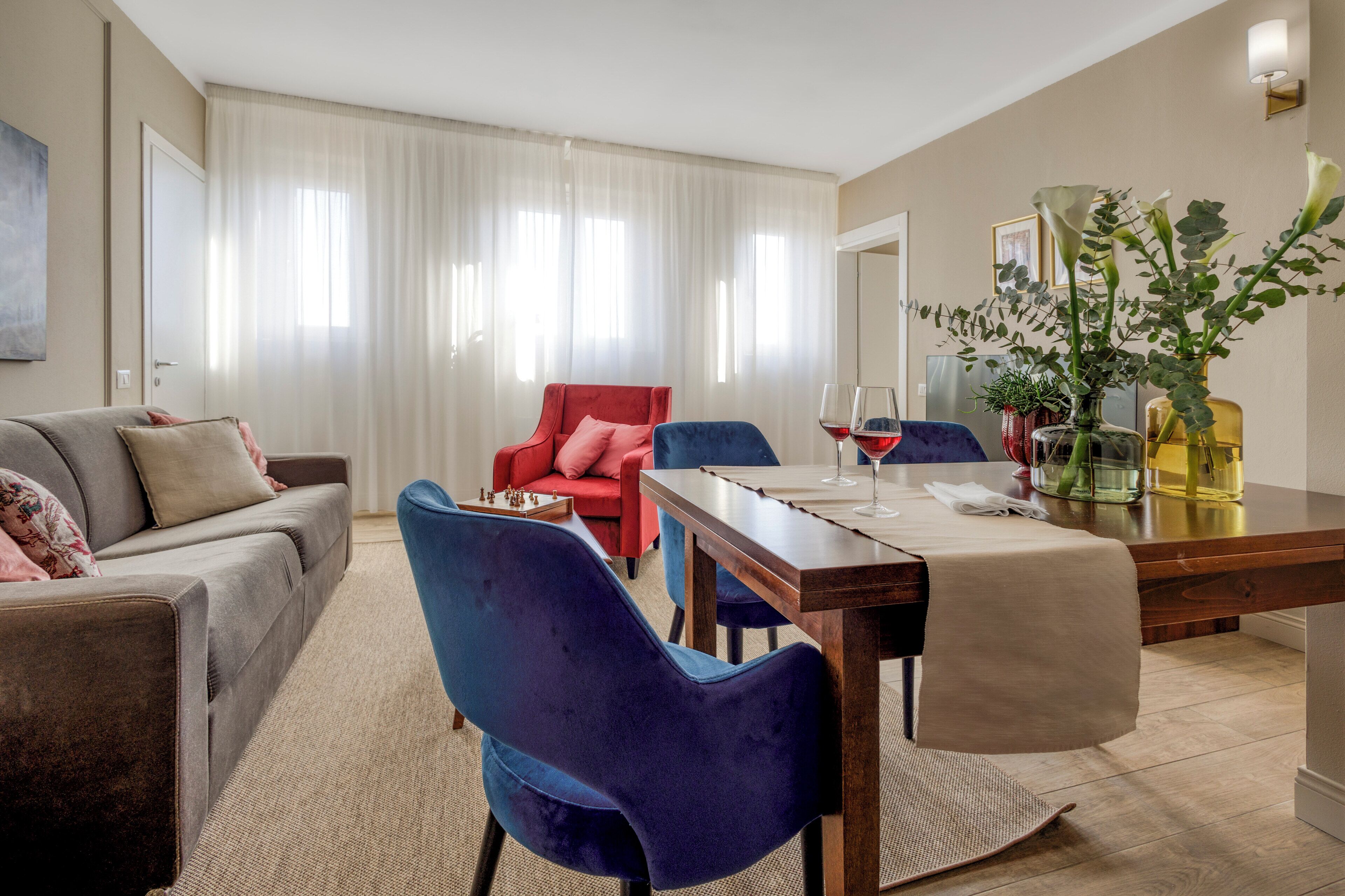 A modern living room with a sofa, a red chair, a dining table with chairs, and a vase with flowers.