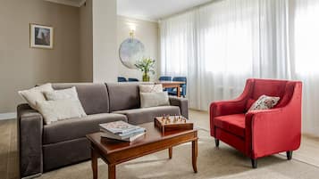Una sala de estar con un sofá, un sillón rojo, una mesa de centro con revistas y un tablero de ajedrez.