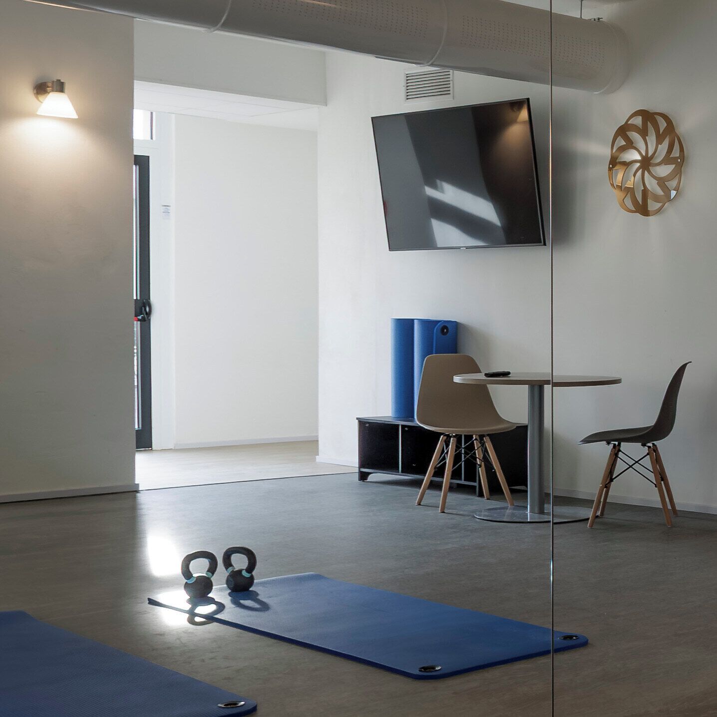 Habitación moderna con televisor grande, colchoneta de ejercicios azul, mancuernas y una pequeña mesa con sillas.
