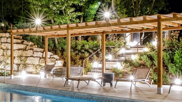 An outdoor swimming pool with a wooden pergola, lounge chairs, and a stone wall backdrop. - Guild Living Siena Senior Resort & Residence (Siena)