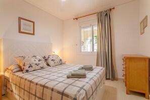 Un dormitorio con cama, almohadas, mesita de noche, armario de madera, ventana con cortinas e imagen enmarcadas en la pared.