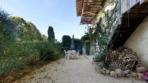 Un chemin de gravier mène à une table et des chaises blanches installées à l’extérieur, avec une pile de bois de chauffage sur la droite et un feuillage dense de chaque côté.