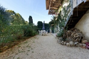 A gravel path leading to a white table and chairs set up outdoors, with a pile of firewood to the right and dense foliage on either side.