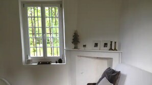 Een woonkamer met een witte bank, een bijzettafeltje met een boek, een decoratief vloerkleed, een open haard en een raam met uitzicht op groen.