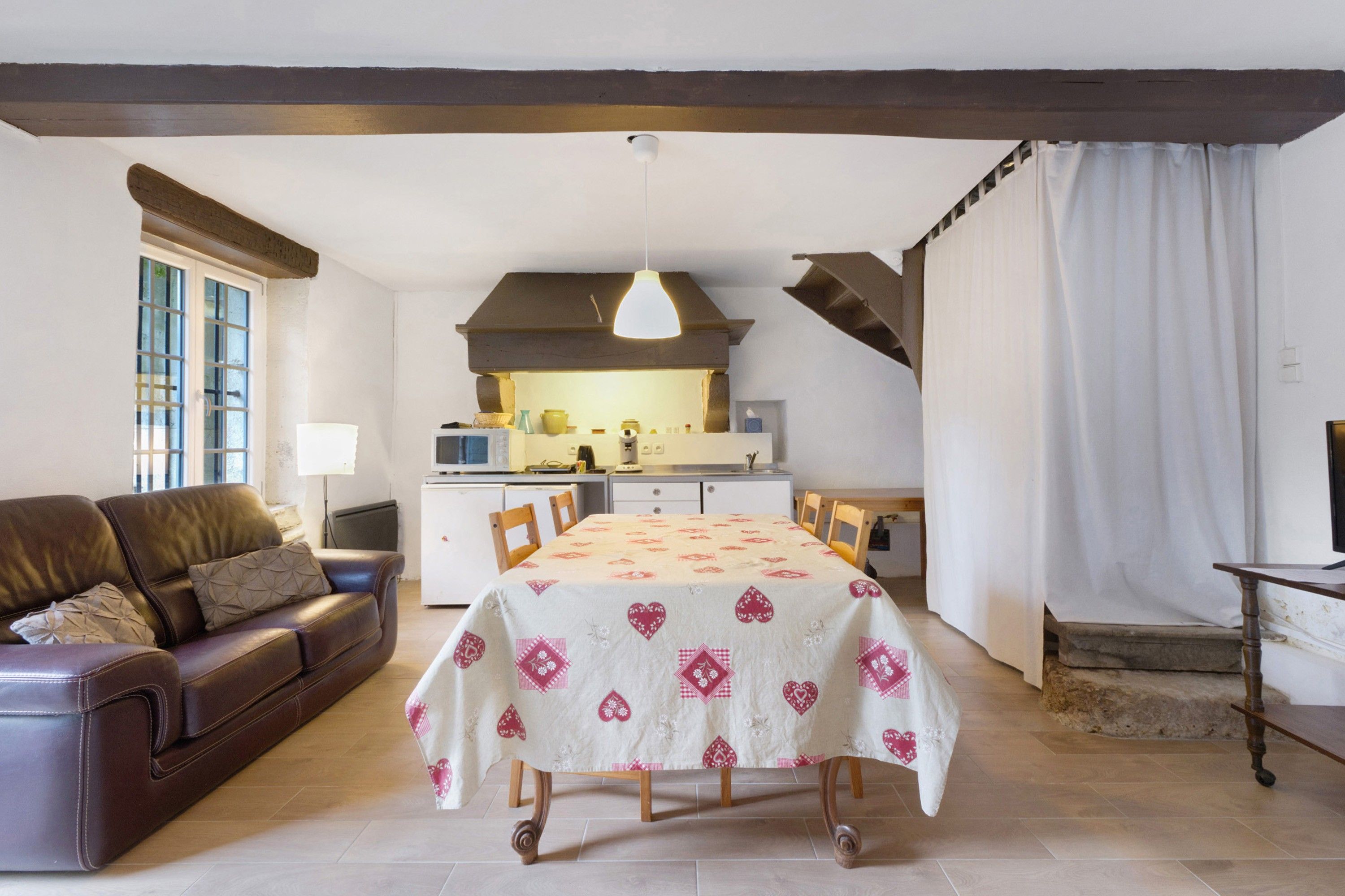 Uma sala de estar acolhedora com sofá, mesa de jantar com toalha de mesa estampada e área de cozinha ao fundo.