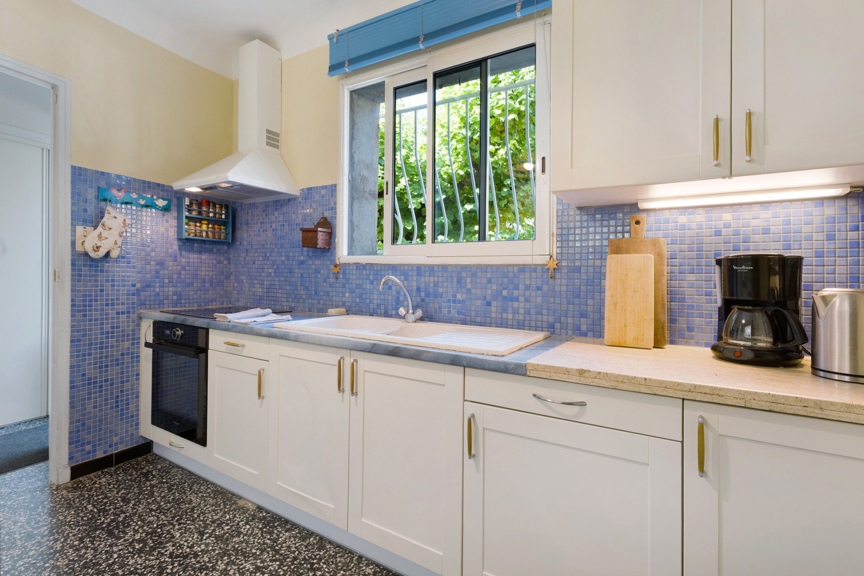 Een keuken met witte kastjes, een blauwe betegelde achterwand, een raam met beplanting buiten, een spoelbak en een koffiezetapparaat.