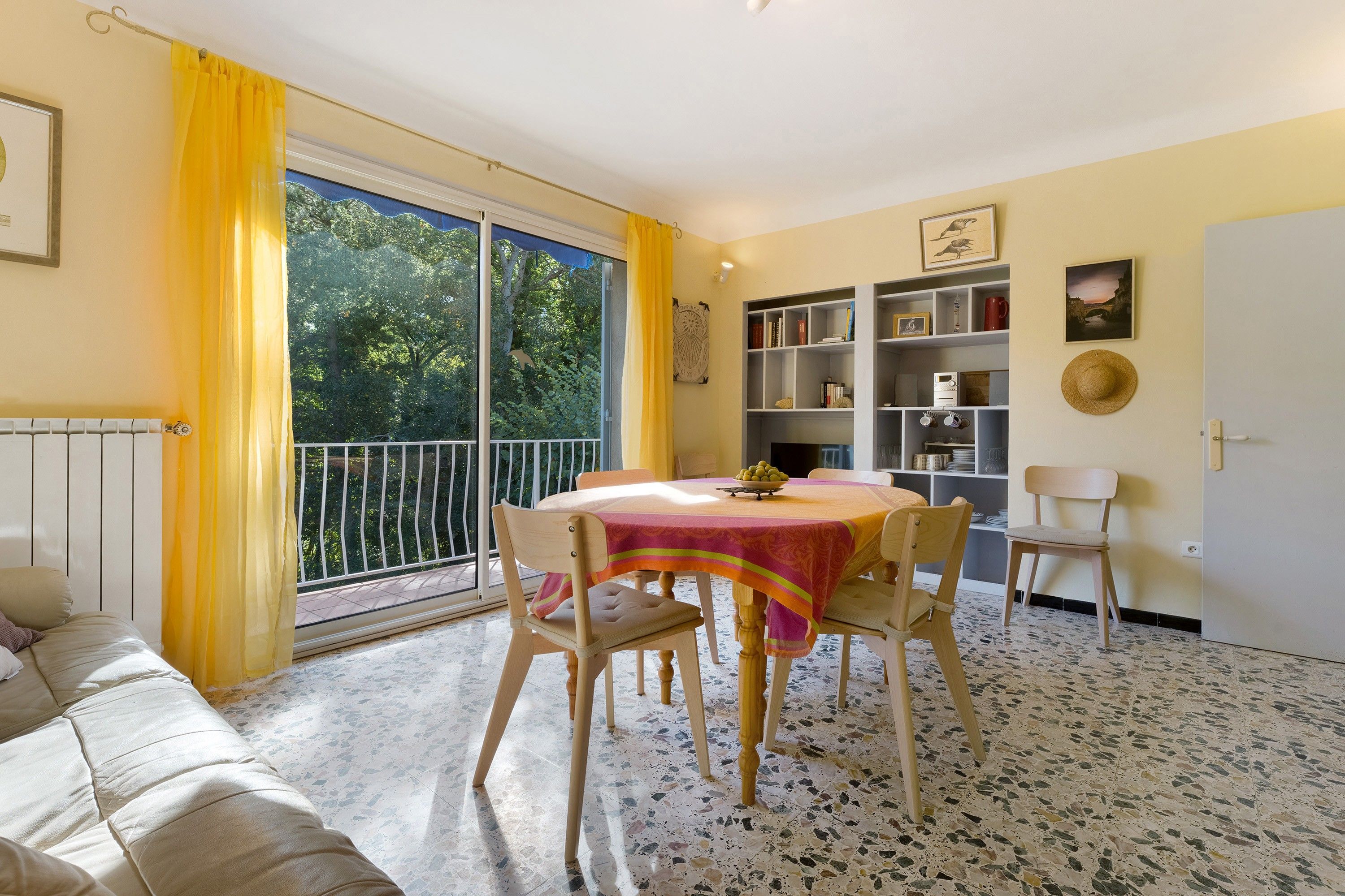 A room with a dining table, chairs, a sofa, a bookshelf, and a window with curtains.