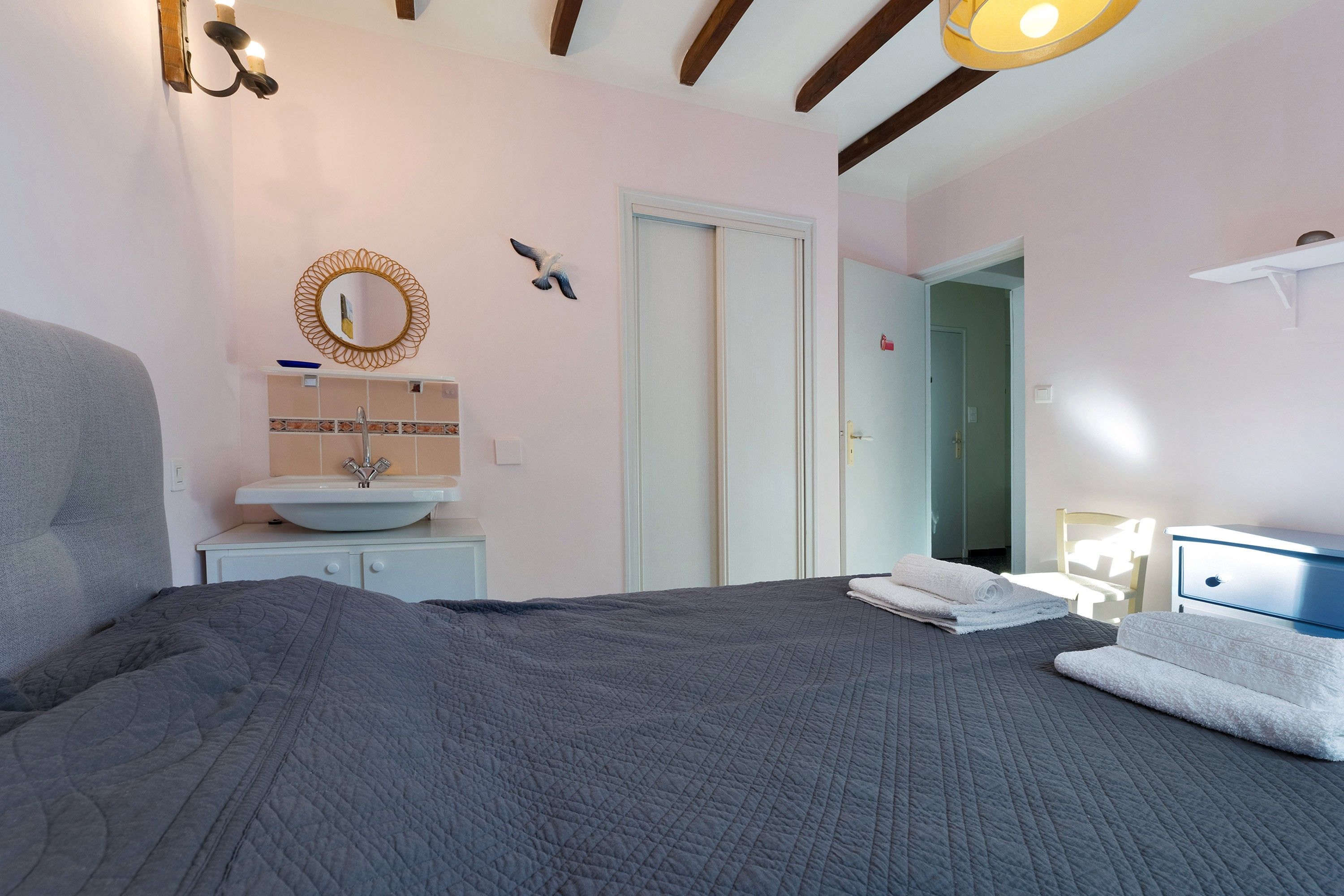 A bedroom with a large bed, a bedside table, a mirror, a sink, and a door leading to another room.
