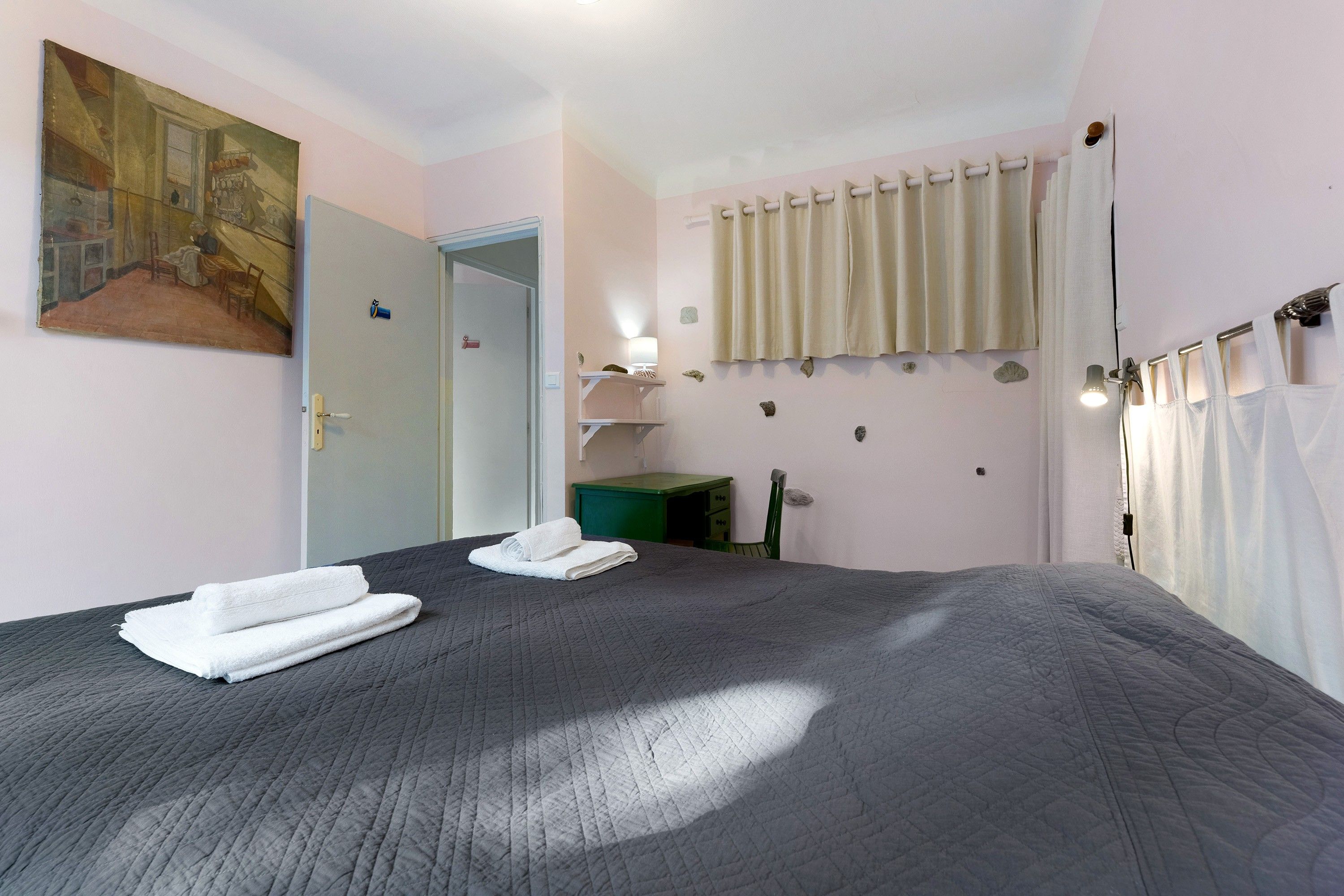 A bedroom with a large bed, a painting on the wall, a door, a small table, and a window with curtains.