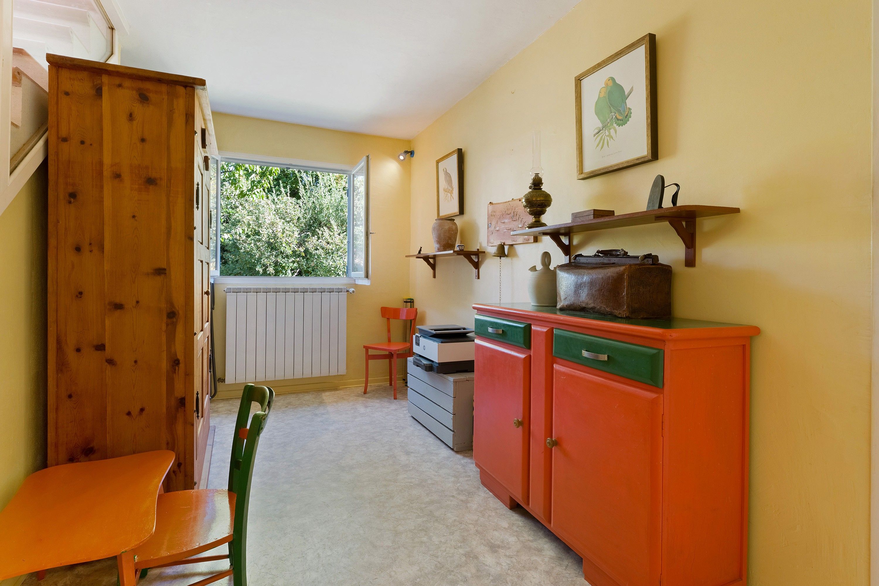A room with a wooden wardrobe, a red cabinet, a chair, a radiator, and a window with a view of greenery.