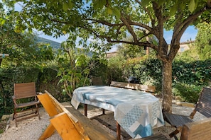 Un espace extérieur aménagé avec une table et des bancs sous un arbre, entouré de verdure et offrant une vue sur une montagne.