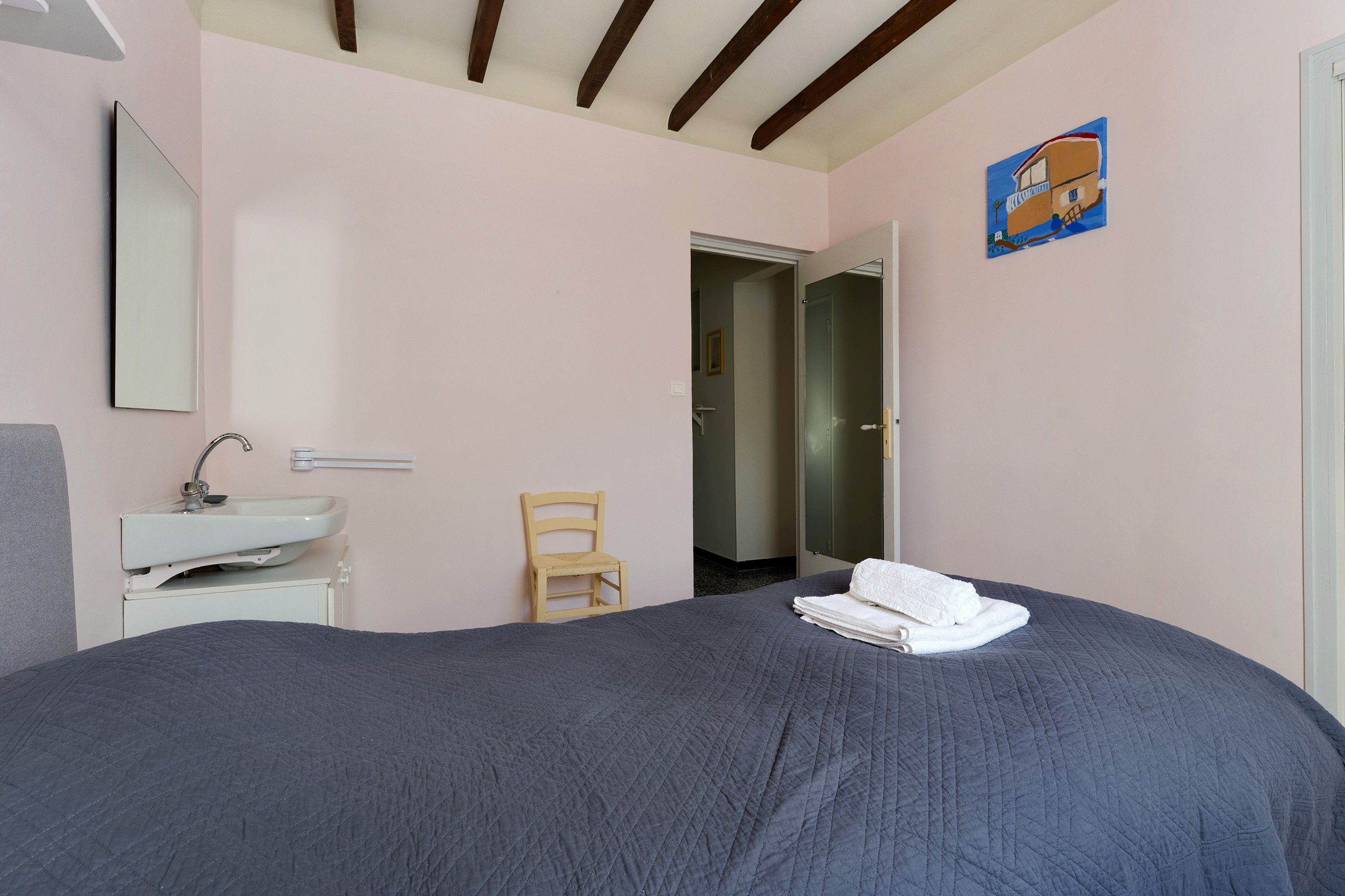 Un dormitorio con cama, mesita de noche, una silla, un espejo y una puerta que da a otra habitación.