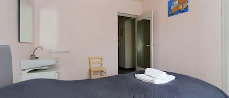 Un dormitorio con cama, mesita de noche, una silla, un espejo y una puerta que da a otra habitación.