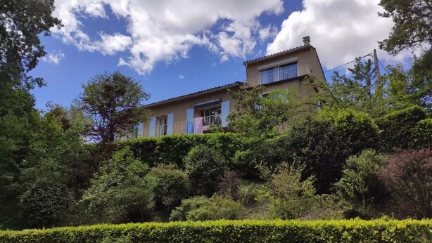 Ein zweistöckiges Haus mit Balkon, umgeben von üppiger Vegetation und blauem Himmel.