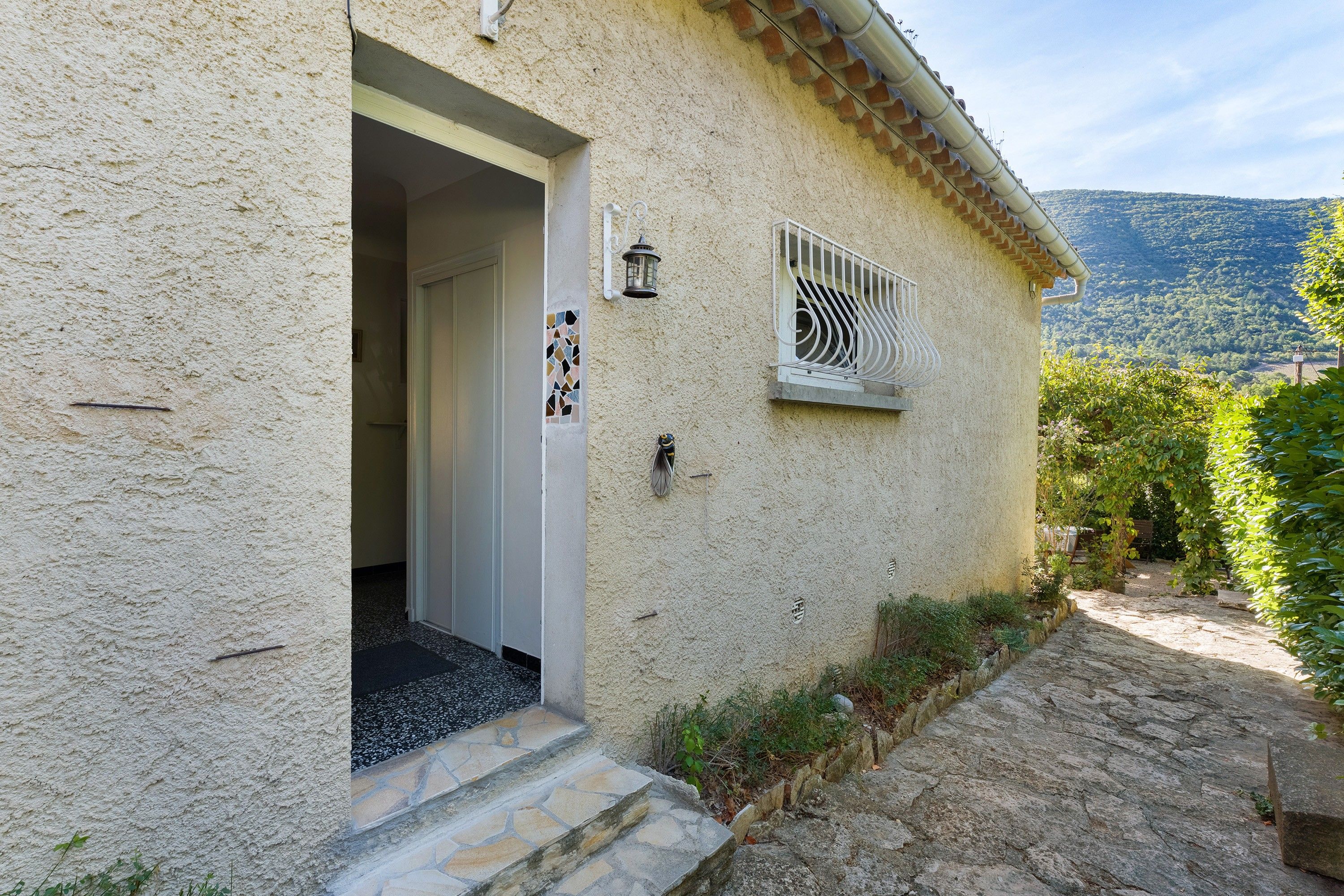 A rustic building with a stone staircase leading to a door, a small window with a metal grill, and a view of a mountainous landscape.