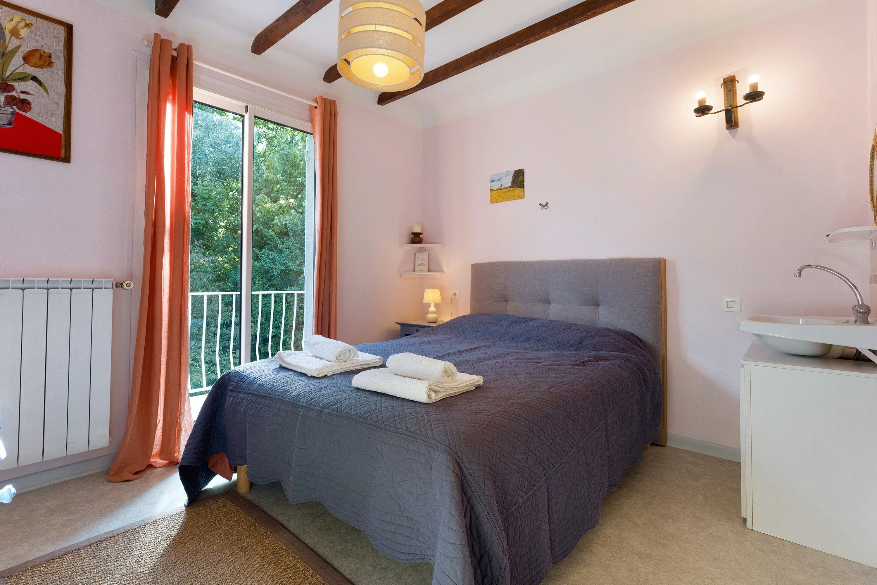 A bedroom with a bed, a nightstand, a window with curtains, a radiator, and a wall-mounted light fixture.