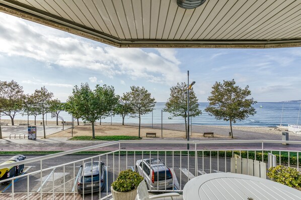 Ein Balkon mit Meerblick, ein Parkplatz mit Autos und ein Strand mit Bäumen und Bänken.