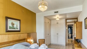 Une chambre d’hôtel avec un lit, une tête de lit en bois, un lustre, un tableau encadré au mur, une chaise, une armoire et une salle de bain visible à travers une porte ouverte.