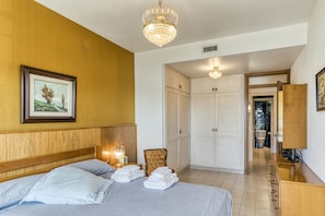 Habitación de hotel con cama, cabecera de madera, candelabro, un cuadro en la pared, una silla, armario y baño visible a través de una puerta abierta.