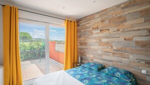 Une chambre dotée d’une grande porte-fenêtre coulissante donnant sur un balcon, d’un lit recouvert d’une couvre-lit aux motifs bleus et verts, et d’un lambris en bois.