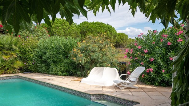 Une piscine dont le bord est recouvert de carrelage, entourée d’une végétation luxuriante et agrémentée d’un fauteuil de relaxation blanc.