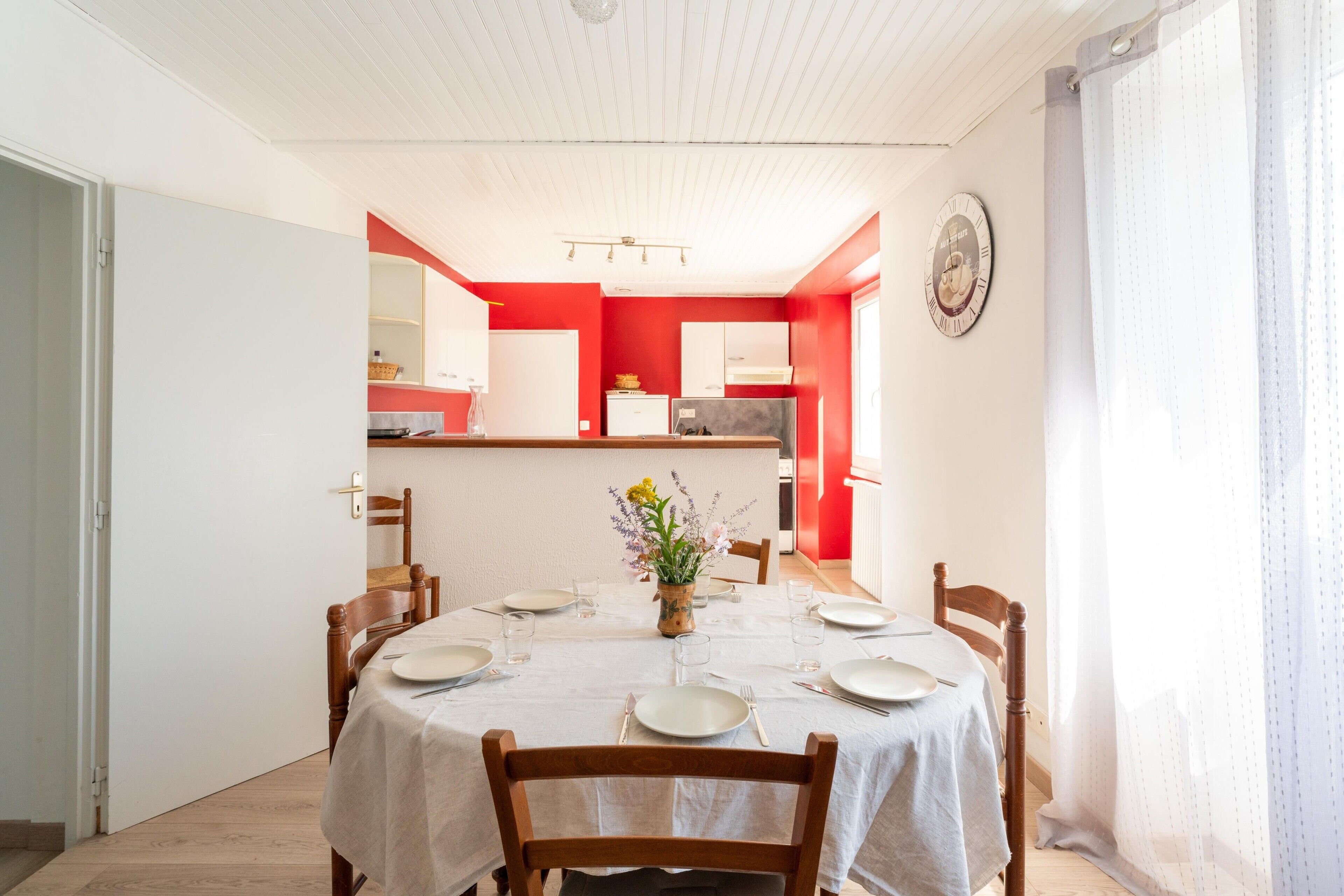 A dining area with a table set for six, a kitchen in the background, and a clock on the wall.