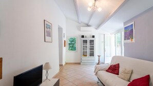 A living room with a TV, a sofa with red cushions, a lamp, and a window with a view of greenery. - Holiday Home "Gîte Lou Barry" with Shared Pool & Wi-Fi (Mirabel-aux-Baronnies)