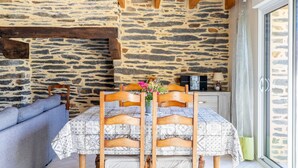 Un coin repas avec un mur en pierre, des poutres en bois, une table recouverte d’une nappe à motifs et un canapé.