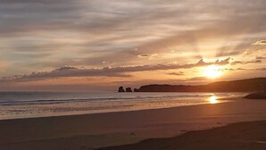 Una playa con atardecer, mar tranquilo y formaciones rocosas a lo lejos.
