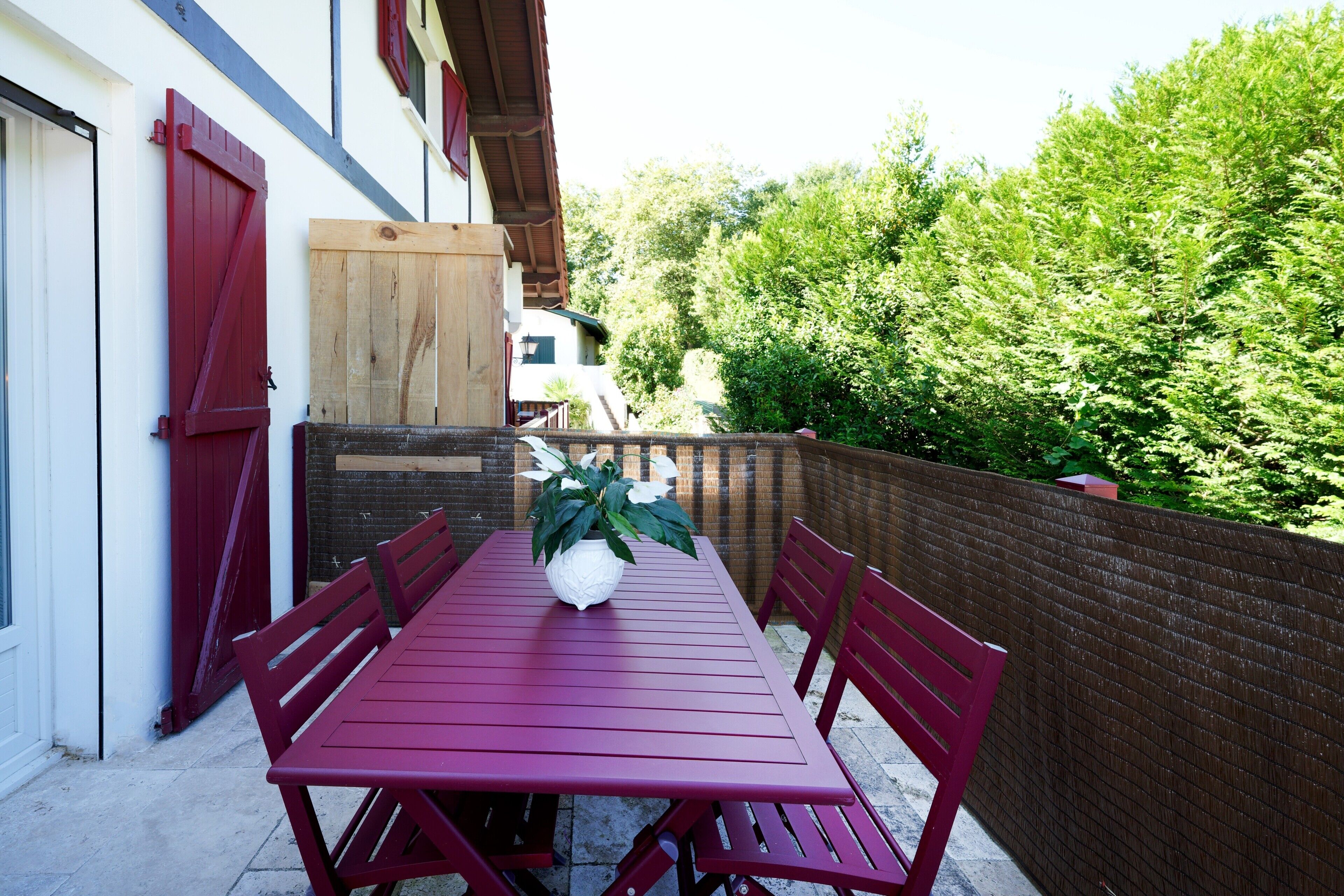 Une table en bois rouge, entourée de chaises, un pot de fleurs et une porte en bois dans une cour bordée d’un mur de briques et de verdure.