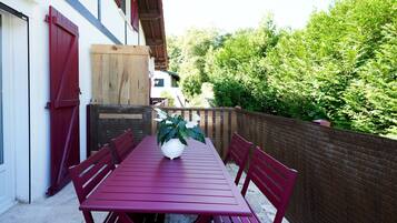 Une table en bois rouge, entourée de chaises, un pot de fleurs et une porte en bois dans une cour bordée d’un mur de briques et de verdure.