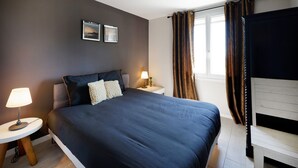 Un dormitorio con cama, mesitas de noche, lámpara, ventana con cortinas e imagen colgada en la pared.