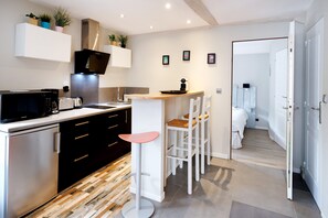 A modern kitchen with a microwave, toaster, and coffee maker, a white bar stool, and a view into a bedroom.