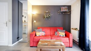 A living room with a red sofa, a wooden coffee table, a wall-mounted light fixture, and a decorative hanging piece. - Holiday Apartment "Escapade Au Pays Basque 2 to 4 Guests" with Balcony, Shared Garden & Wi-Fi (Urrugne)