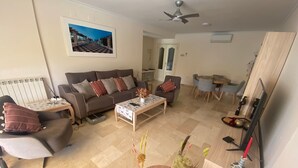 A living room with a sofa, coffee table, and a TV. There is a dining area with chairs and a table. A ceiling fan is visible. - Apartment 'Apartamento Cal Albir' with Shared Pool, Wi-Fi and Air Conditioning (l'Alfàs del Pi)