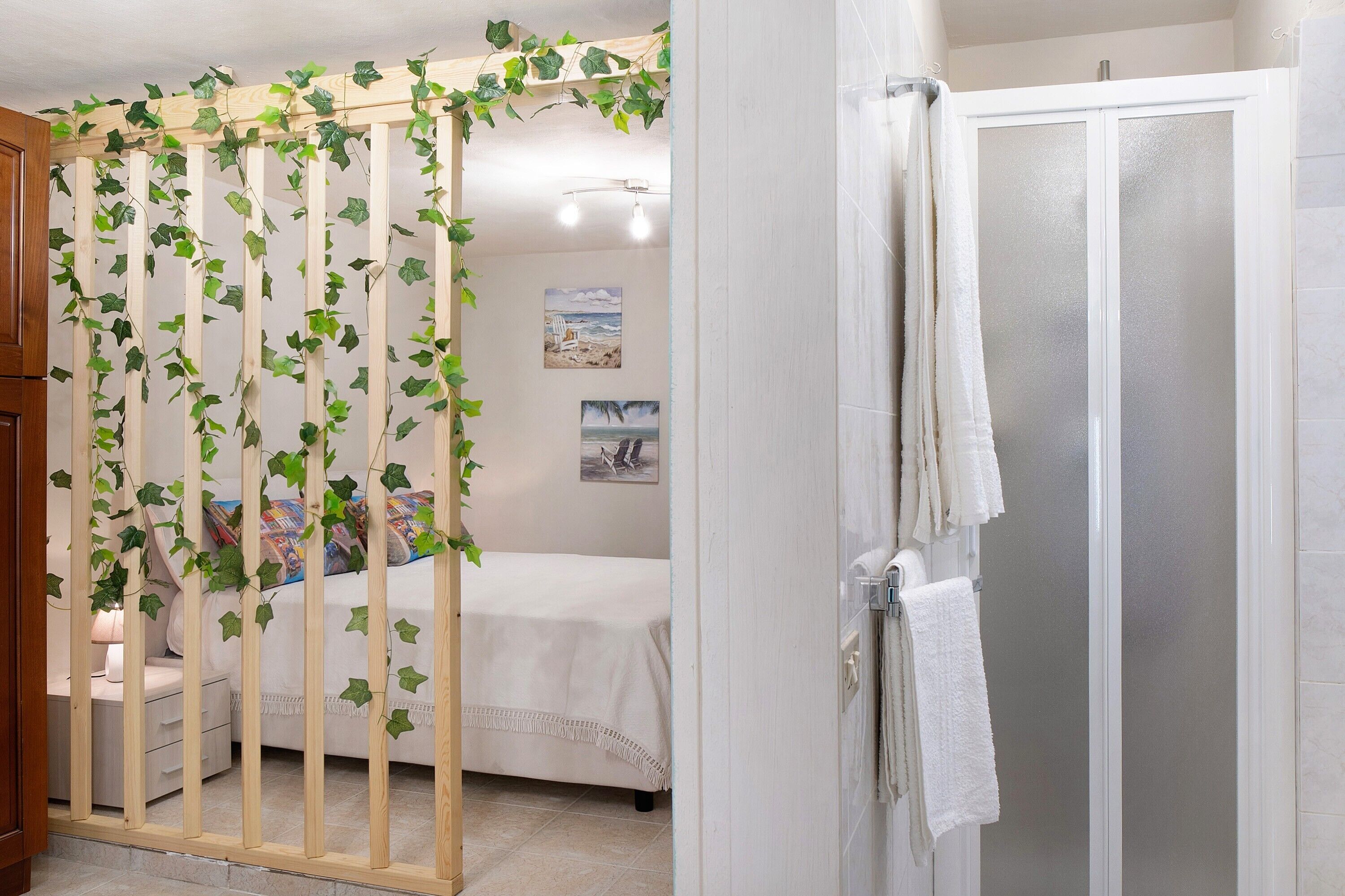 Un dormitorio con cama, un separador de madera con plantas, un baño con ducha y una pared con cuadros.