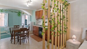 A kitchen with wooden cabinets, a dining table with chairs, a wooden partition with greenery, and a bedside table with a lamp.