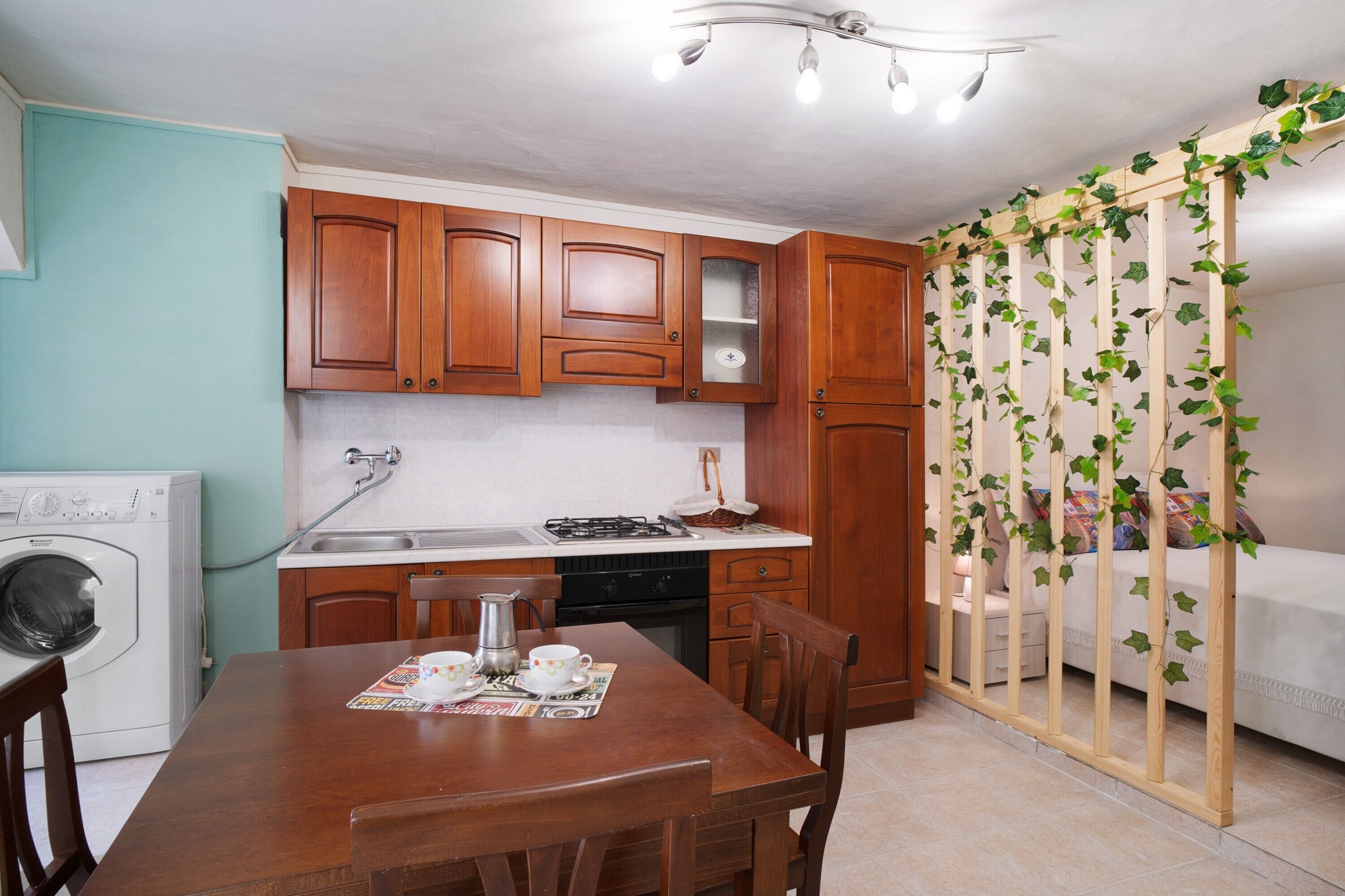 Una cucina funzionale con mobili in legno, una lavatrice, un tavolo da pranzo con sedie e un letto separato da un pannello in legno.